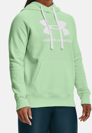 Person iført lysegrøn Under Armour hættetrøje med hvide snore og logo, kombineret med mørkeblå bukser.