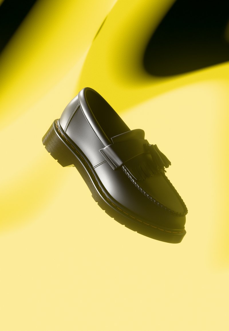 Mocassins de couro preto com um tassel decorativo, apresentando uma borda costurada e sola grossa e texturizada. Colocados sobre um fundo amarelo.