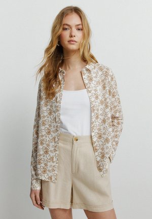 Młoda kobieta o długich blond włosach, ubrana w biały top na ramiączkach, beżowe krótkie spodenki z wysokim stanem oraz rozpiętą kwiecistą koszulę na guziki.