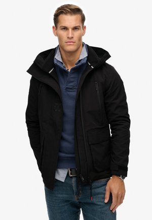 Chaqueta impermeable negra con capucha, cierre frontal con cremallera, múltiples bolsillos y un acabado texturizado. Llevada sobre una camisa azul y jeans azules.