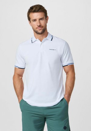 Hombre vistiendo una camiseta tipo polo blanca con ribete azul marino y logo "Hackett", pantalones verdes, de pie con las manos en los bolsillos contra un fondo liso.