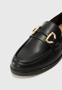 Juodas odinis loafers su aukso detalėmis, su lygiu paviršiumi, apvaliu pirštu ir siuvinėta detale palei padą.