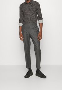Chemise noire à motifs avec boutons et manches retroussées, associée à un pantalon gris ajusté et des baskets noires à lacets.