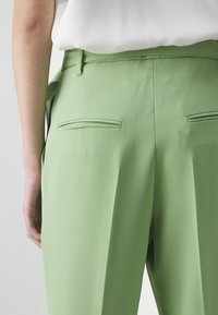 Gros plan d'une personne portant un pantalon vert clair sur mesure avec des poches passepoilées à l'arrière et un haut blanc rentré.