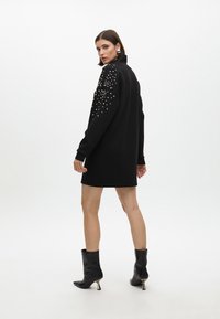 Schwarzes, übergroßes Kleid mit langen Ärmeln, verziert mit silbernen Nieten an einer Schulter. Kombiniert mit schwarzen Stiefeletten und einem hohen Kragen.