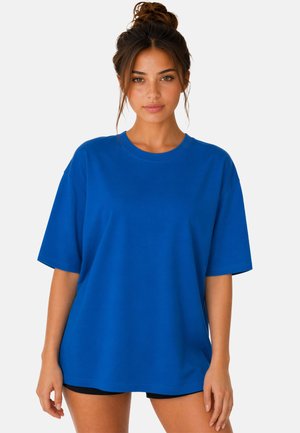 Femme aux cheveux bruns en chignon portant un t-shirt bleu oversize à manches courtes et un short noir, debout sur un fond blanc.