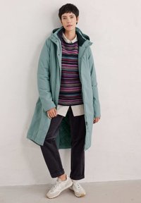 Lange teal pufferjas met een capuchon, gelaagd over een meerkleurige gestreepte trui, wit shirt en donkere broek, gecombineerd met lichte sneakers.