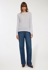 Maglione a maniche lunghe grigio chiaro, girocollo, abbinato a jeans blu in denim larghi e scarpe marroni a punta, in piedi contro uno sfondo chiaro.