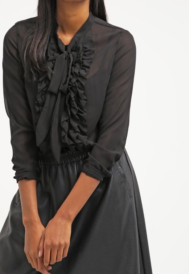Blouse noire transparente avec devant à volants et détail de col noué, manches longues et poignets ajustés. Portée avec une jupe noire, texture lisse.