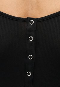 Μαύρο ριμπ τοπ με στρογγυλή λαιμόκοψη και έξι ασημένια κουμπώματα τύπου σναπ στην πλάτη, που αναδεικνύει υφή πλεκτού υφάσματος.