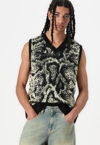 Gebreide mouwloze trui in groen en zwart dierenprint, met een V-halslijn, ribgebreide randen en een ontspannen pasvorm. Draagt over lichte spijkerbroek.