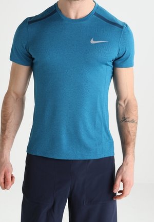 Mand iført en blå kortærmet sportstrøje med et hvidt Nike-logo og mørke shorts, står med afslappede arme.