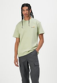 Light green cotton t-shirt with short sleeves and a round neck. Features a small embroidered logo in orange. Paired with dark gray cargo pants.