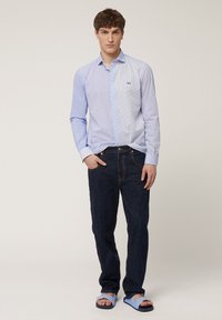 Lichtblauw overhemd met knopen en een stippenpatroon, lange mouwen, gecombineerd met donkere jeans en gestreepte sandalen. Opmerkelijk logo op het shirt.