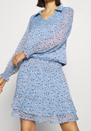 Femme portant un chemisier bleu à manches longues et une jupe plissée assortie avec de petits motifs abstraits en forme de cœur et un ourlet à volants.