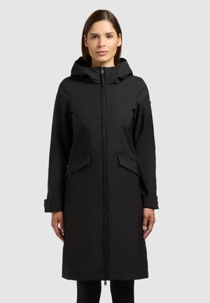 Femme portant un manteau noir à capuche longueur genou avec fermeture éclair frontale et poches à rabat, debout devant un fond clair uni.