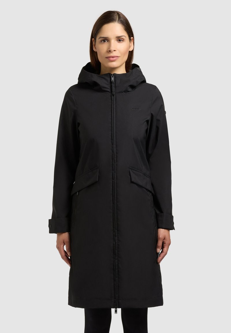 Kobieta ubrana w czarny, sięgający kolan płaszcz z kapturem, z przednim zamkiem błyskawicznym i kieszeniami z klapkami, stojąca na jasnym, jednolitym tle.