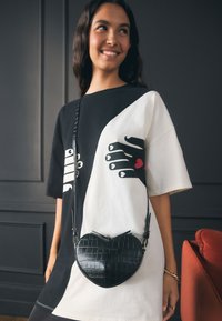 Melna mākslīgās krokodila ādas somiņa sirds formas ar plānu siksnu, saskaņota ar melnu un baltu pārmērizmēra grafisko T-kreklu ar roku zīmējumiem.