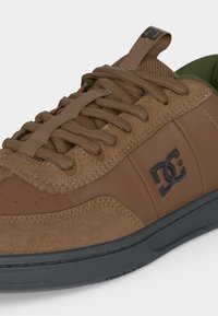 Gros plan d'une basket marron avec des détails en daim et cuir, des lacets marron, un logo DC noir sur le côté et une semelle gris foncé.