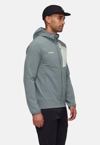 Lys grå jakke med hætte, frontlynlås og brystlomme. Tekstil med struktureret overflade og kontrastfarvet off-white panel; har subtile stikninger.