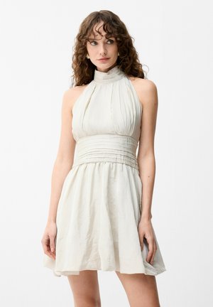 Mujer con cabello rizado que lleva un vestido sin mangas, de cuello alto y color crema, con una cinturilla ajustada y plisada.