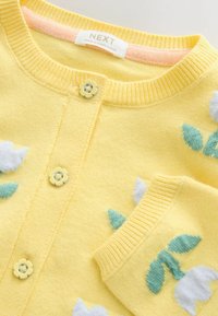 Dzeltena adīta jaka ar baltu un zaļu ziedu rakstiem, apaļām pogām zieda formā un ribota apkakle un aproces.