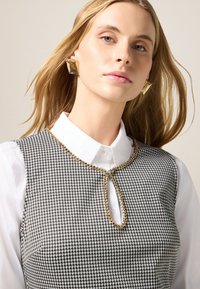 Schwarz-weiße Hahnentrittmuster-Weste mit einem Schlüsselloch-Ausschnitt, goldener Kettendetail und weißem Hemd darunter. Glattes Haar, natürliches Make-up.