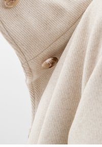 Béžová vlněná tkanina s jemnou strukturou, která má šikmý rib vzor a velké průhledné knoflíky na jedné straně.