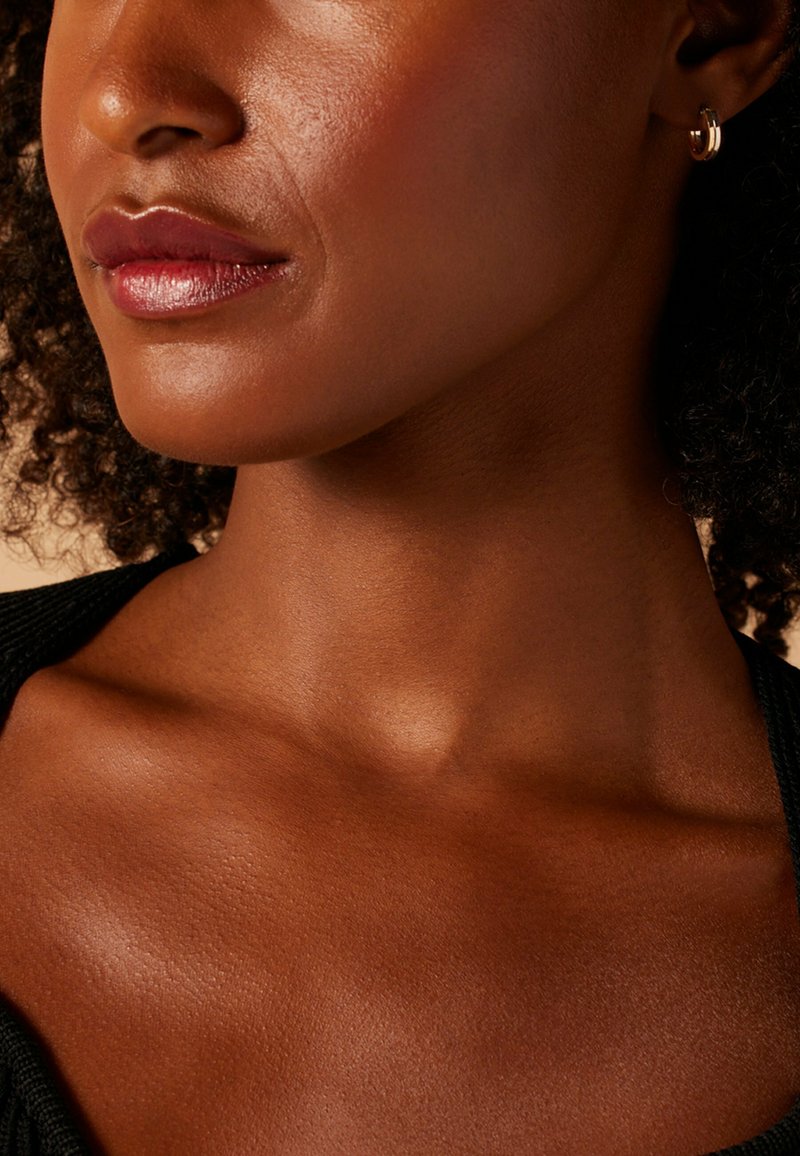 Close-up van een nek en gezicht met een gladde, mediumbruine huid, subtiele make-up en een kleine gouden creool. Donker, krullend haar is gedeeltelijk zichtbaar.