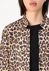 Jachetă cu imprimeu leopard realizată dintr-un material texturat, cu un design fără guler și deschis la front, purtată peste un top negru.