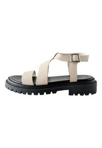 Sandale din piele bej cu două barete frontale și cataramă reglabilă la gleznă, având o talpă groasă din cauciuc negru cu model texturat.