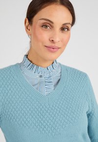 Maglione a maglia azzurro chiaro con un motivo testurizzato, design a scollo a V, sovrapposto a una camicia a righe bianca e azzurro. 
