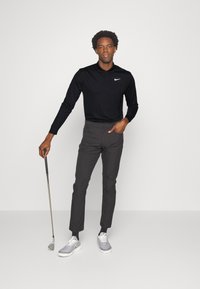 Crna majica s dugim rukavima i suptilnim Nike logotipom; sivi lagani golf hlače; drži golf štap; sivi sportski tenisice s bijelim detaljima.