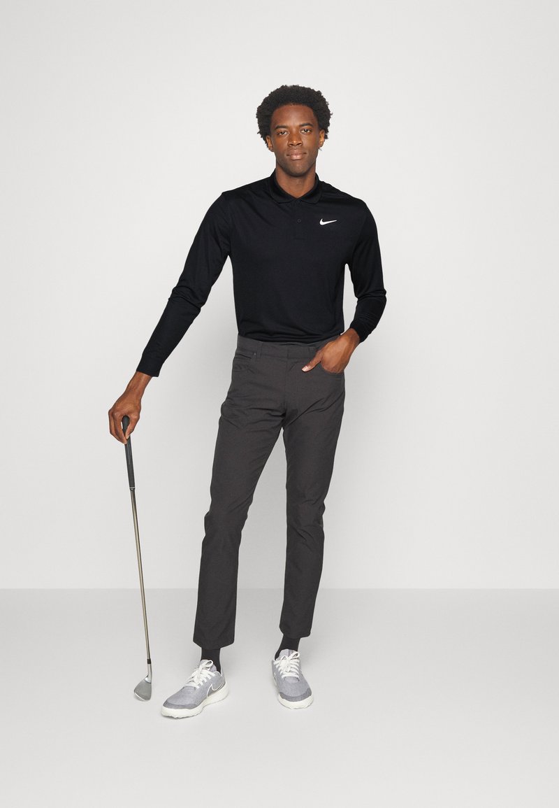 Polo shirt a maniche lunghe nera con logo Nike discreto; pantaloni da golf leggeri grigi; tiene in mano una mazza da golf; scarpe sportive grigie con dettagli bianchi.
