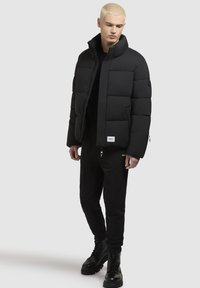 Schwarze Daunenjacke mit gesteppten Paneelen, hohem Kragen und Logodetails. Trägt man über einem schwarzen Shirt und Jogginghosen, kombiniert mit schwarzen Schnürstiefeln.
