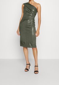 En axelbandslös midiklänning i metalliskt olivgrönt med rynkad textur, tight silhuett och knälång nederdel. Öppna svarta klackar.
