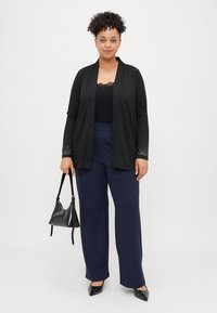 Μαύρο κασκόλ με υφασμάτινα μανίκια, φορεμένο πάνω από μια τοπ με δαντελένια περικοπή; συνδυασμένο με ναυτικές παντελόνες wide-leg και μαύρες στιλέτο με καρφιά, κρατώντας μια μικρή τσάντα.