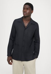 Tmavomodrá košeľa s dlhým rukávom, golierom, prednými gombíkmi, dvoma vreckami a uvoľneným strihom, vyrobená z textúrovanej tkaniny.
