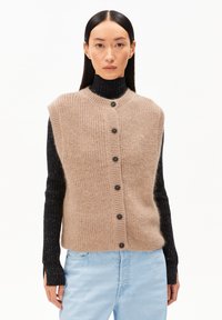 Beige gebreid vest met een ronde hals, vijf zwarte knopen aan de voorkant, gedragen over een donker geribd shirt met lange mouwen. Lichtblauwe spijkerbroek.