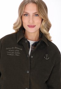Ciemnozielona kurtka sztruksowa z zapięciem na guziki, ozdobiona haftowanym napisem i emblematem kotwicy. Posiada karciane podszewki i klasyczny kołnierz.
