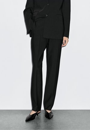 Black tailored suit featuring a satin finish, structured shoulders, and straight pants. Accompanied by pointed, open-back black heels.