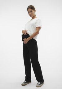 Calças de maternidade pretas combinadas com uma t-shirt branca, mostrando uma barriga redonda, estilizadas com sapatilhas desportivas pretas e cinzentas. Tecido suave, corte descontraído.