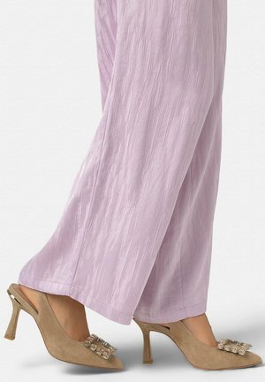 Tacones beige de gamuza con tira trasera y hebillas decoradas con joyas, combinados con pantalones anchos texturizados de color púrpura claro.