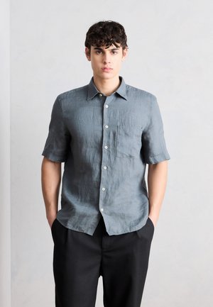 Giovane uomo con una camicia grigia a maniche corte abbottonata e pantaloni neri, in piedi con le mani nelle tasche davanti a uno sfondo chiaro e uniforme.