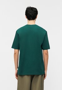 Άτομο με κοντά σκούρα μαλλιά που φοράει απλό σκούρο πράσινο κοντομάνικο πουκάμισο και μπεζ παντελόνι, απεικονιζόμενο από πίσω σε λευκό φόντο.