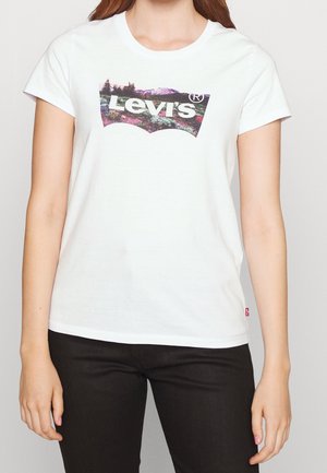 Mujer con una camiseta blanca de Levi's con un logo estampado de paisaje y pantalones negros, de pie contra un fondo claro y liso.
