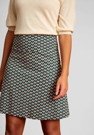 Kvinde iført en beige kortærmet top og et højtaljet A-formet skørt med et flerfarvet geometrisk mønster.