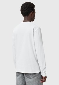 Weißes Langarmshirt mit rundem Halsausschnitt. Weiches Baumwollmaterial, lässige Passform und Standardmanschetten. Kombiniert mit hellgrauen Jeanshosen.