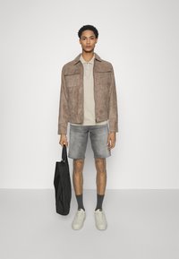Giacca in suede marrone, polo beige, pantaloni corti in denim grigi e sneakers bianche. Tiene una borsa tote nera, in piedi contro uno sfondo neutro.