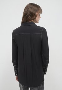 Camicia nera con maniche lunghe, cuciture bianche, dettaglio di piega posteriore e polsini abbottonati. Il tessuto sembra leggero con una texture liscia.